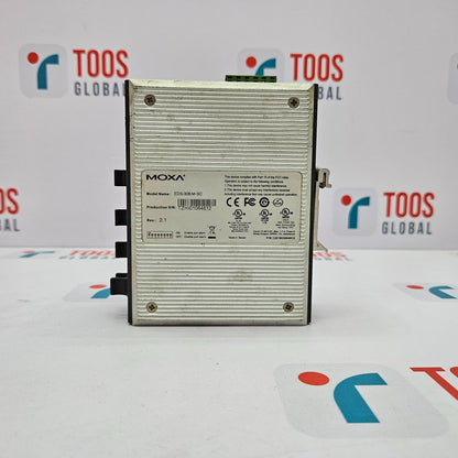 A MOXA EDS-308-M-SC 8-Port Unmanaged Ethernet Switch, with a silver body and black ports, positioned vertically on a table with a logo in the background.
