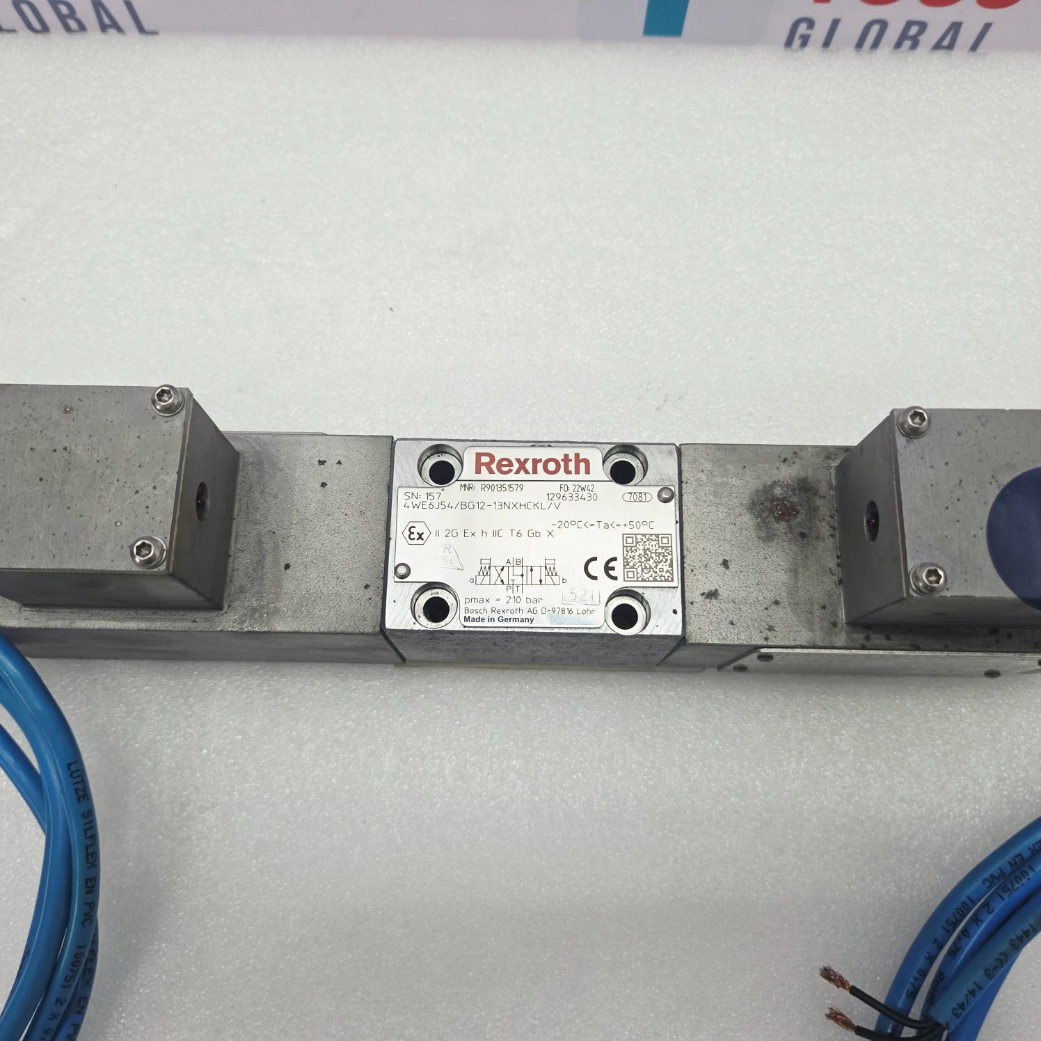 A Rexroth industrial pneumatic valve component with a silver metal plate featuring the red Rexroth logo, mounted on a gray metal bar. Blue pneumatic wires are visible in the foreground on a white background.